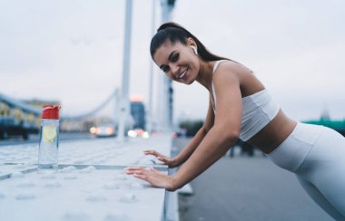 Kulaklıklı, gülümseyen, müzik dinleyen ve briçe yaslanıp şehirde spor yaparken kameraya bakarak şınav çeken sporcu kadının yan görüntüsü.