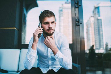 Resmi olarak odaklanmış sakallı genç işadamı modern kafe terasında oturuyor ve cep telefonuyla konuşuyor.