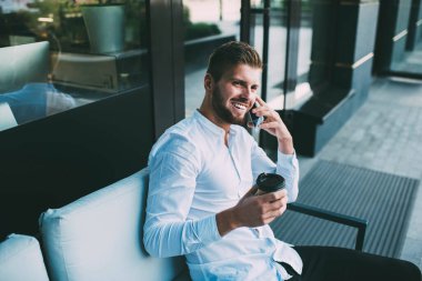 Yukarıdan, dişlek tebessümlü neşeli bir erkeğin elinde bir fincan kahveyle caddede otururken gözlerini kaçırması ve telefon görüşmesi yapması.