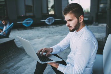 Konsantre olmuş sakallı iş adamının modern netbook 'ta resmi tişört giyip sokak kafesinde rahat bir koltukta otururken görüntüsü.