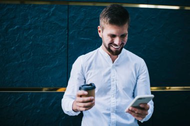 Beyaz tişörtlü, mutlu sakallı adam, gülümseyip akıllı telefon ekranına bakarken kahve molası verip caddeye çıkıyor.