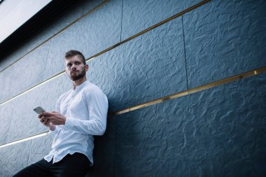 Beyaz tişörtlü, düşünceli, sakallı bir erkeğin modern binanın taş duvarına yaslanırken gözlerini kaçırıp akıllı telefonuna bakması.