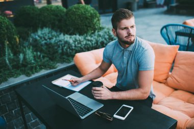 Kendine güveni yüksek, serbest çalışan rahat bir genç adam kanepede bilgisayar başında çalışıyor ve terasta kahve içerken cep telefonunun yanındaki masada not defterine notlar yazıyor.
