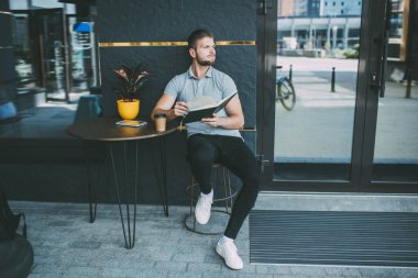 Sıradan kıyafetli, üzgün sakallı bir erkek sokağın tahtadan masasına oturmuş kitap okurken gözlerini kaçırıyor.
