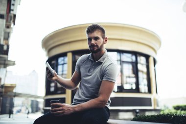 Çok ciddi sakallı bir erkek cep telefonu kullanıyor ve sokaktaki kentsel binanın bulanık arka planında kameraya bakıyor.