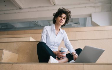 Milenyum Kafkas kızı dizüstü bilgisayar kullanırken gözlerini kaçırıyor. Modern başarılı kadın kavramı. Kahveli genç kıvırcık kadın çalışma odasındaki ahşap bankta oturuyor.
