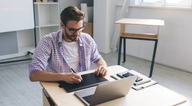Milenyum Kafkas işadamı laptoptan izliyor ve grafik tabletine bir şeyler yazıyor. Modern başarılı adam kavramı. Gözlüklü genç adam açık uzay ofisinde masada oturuyor.