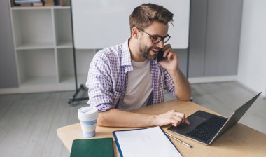 Milenyum Kafkas işadamı cep telefonuyla konuşuyor ve dizüstü bilgisayardan bir şeyler izliyor. Modern başarılı adam kavramı. Gözlüklü genç adam açık uzay ofisinde masada oturuyor.