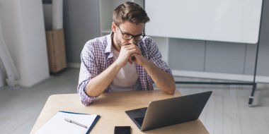 Milenyum Avrupalı işadamı dizüstü bilgisayardan bir şey izliyor ve bir şeyler düşünüyor. Modern başarılı adam kavramı. Açık uzay ofisindeki masada oturan gözlüklü genç adam.