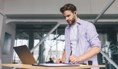 Milenyum Kafkas işadamı laptoptan izliyor ve grafik tabletine bir şeyler yazıyor. Modern başarılı adam kavramı. Ofisteki kablosuz kulaklıklı genç sakallı adam.