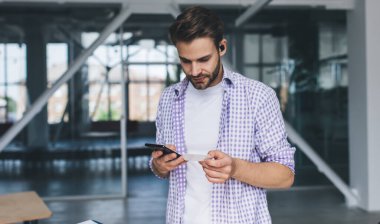 Milenyum Kafkas işadamı kartvizitten bir şey arıyor ve akıllı telefondan yazıyor. Modern başarılı adam kavramı. Açık uzay ofisindeki kablosuz kulaklıklı genç adam.
