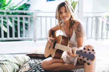 Otelin terasında akustik gitar çalan beyaz bir kadın. Eğlence, eğlence ve hobi anlayışı. Tatil ve hafta sonu. Dövmeli gülümseyen kız halıya oturur ve kameraya bakar.