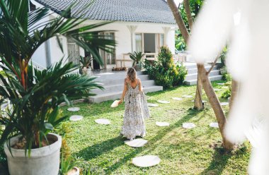 Güneşli bir günde otelin bahçesine çıkan bir kadının arka görüntüsü. Turizm, tatil ve hafta sonu. Deniz kabuğu tutan elbise giyen genç ve güzel bir kız. Bali adasında huzurlu ve huzurlu bir yaşam tarzı