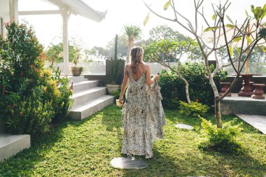 Güneşli bir günde otelin bahçesine çıkan bir kadının arka görüntüsü. Turizm, tatil ve hafta sonu. Deniz kabuğu ve tütsü çubuğu tutan elbise giyen genç bir kız. Bali adasında huzurlu ve huzurlu bir yaşam tarzı