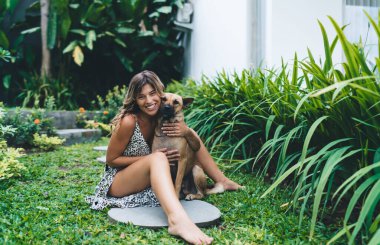Gülümseyen genç bir kadın otel bahçesinde sevimli bir köpeğe sarılıyor. Turizm, tatil ve hafta sonu. Sahibi ve evcil hayvan ilişkisi ve dostluğu. Güzel beyaz kız elbise giyer. Idyllic ve sakin yaşam tarzı