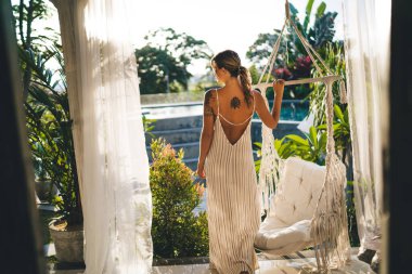 Otelin terasındaki genç bayanın arka görüntüsü. Turizm, tatil ve hafta sonu kavramı. Elbise giyen dövmeli çekici bir kız. Güneşli bir gün. Bali adasında huzurlu ve huzurlu bir yaşam tarzı