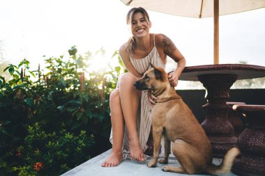 Genç, beyaz bir kız otelin bahçesinde şirin bir köpeği okşuyor. Turizm, tatil ve hafta sonu. Sahibi ve evcil hayvan ilişkisi ve dostluğu. Güneşli bir gün. Bali adasında huzurlu ve huzurlu bir yaşam tarzı
