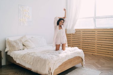 Uzun siyah saçlı, uzun siyah saçlı, kendinden emin genç bayan milenyumun tamamı sabah kollarını kaldırarak yumuşak bir yatakta dizlerinin üzerinde dururken gülümsüyor.