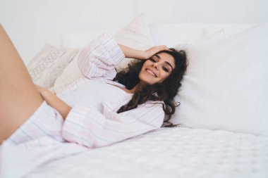 Gece elbiseli mutlu bir kadın başını okşuyor ve evdeki rahat yatak odasında yumuşak bir yatakta dinlenirken kameraya bakıyor.