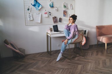 Evin içinde dizüstü bilgisayarı olan, masanın yanında oturan sevimli, genç bir kadın. Tasasız bayan öğrenci modern teknoloji ve modern aletle uzaktan öğrenmenin keyfini çıkarıyor.