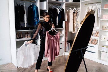 Moda kıyafetleri içinde mutlu bir kadın aynadaki yansımaya bakıyor ve beyaz giysiyi seçiyor dolapta yürüyor ve evinde bir sürü raf var.