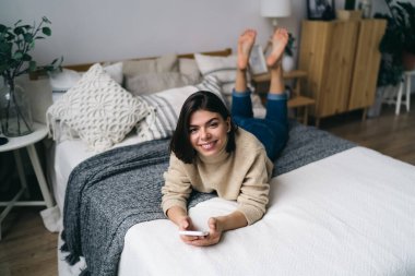 Yukardan gelen kendine güvenen genç İspanyol kadın milenyumu sıcak günlük kıyafetlerle gülümseyen ve kameraya bakan evde rahat bir yatakta uzanan akıllı telefonuyla mesajlaşan.
