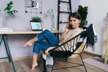 Yan görünüm, koltukta oturan, cep telefonuyla konuşan, evden kameraya bakan, gülümseyen, serbest çalışan bir kadın.