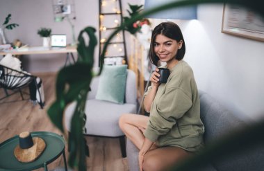 Günlük kıyafetlerle gülümseyen genç bir kadının yan görünüşü. Kanepede oturup kahve içerken dairede kameraya bakması.