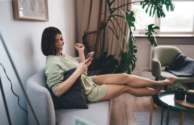 Evdeki oturma odasında rahat bir kanepede dinlenirken günlük kıyafetler giymiş, cep telefonu kullanan neşeli İspanyol kadının yan görüntüsü.