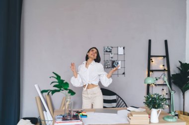 Kulaklıklı, beyaz bluzlu, gülümseyen bir kadın. Ellerini kaldırmış, yeni projeyi düşünüyor ve çağdaş iş yerinde çalışırken başını kaldırıyor.