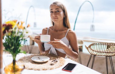 Uzun saçlı mutlu kadın serbest yazar elinde bir fincan latte ile masada oturuyor ve yaz tatilinde tatil köyündeki sahil kafede kameraya bakıyor.