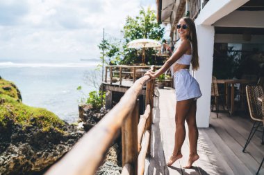 Kenar manzaralı, kendinden emin, uzun saçlı, yaz kıyafetli, güneş gözlüklü genç bir gezgin. Tatil sırasında denizin üzerindeki kayalıklarda duran kafe terasında gülümsüyor.