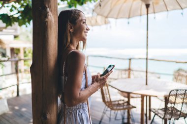 Okyanusa karşı ahşap terasta dikilirken ve yaz tatilinde cep telefonuyla mesajlaşırken gözlerini kaçıran tatmin olmuş genç bir kadın serbest yazar.