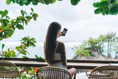 Tanımlanamayan kadın serbest çalışanın, hasır yüksek taburede oturması ve Bali 'de yaz tatilleri boyunca tropik tatil yerlerindeki kafede akıllı telefonlarda kendi portresini çekmesi.