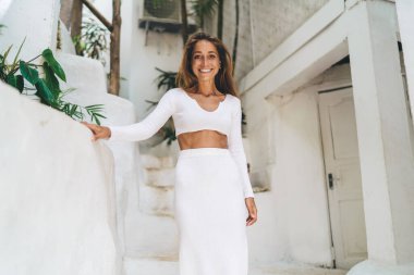 Beyaz etekli, üst üste gülümseyen, kameraya bakan ve yaz tatilinde tropik oteldeki taş duvara yaslanan mutlu kadın serbest yazar.