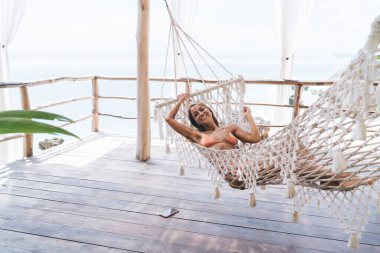 Uzun siyah saçlı, uzun boylu, bikinili, mutlu bir kadın. Güneş ışığı altında, ahşap verandada yatarken kameraya bakıp gülümsüyor.