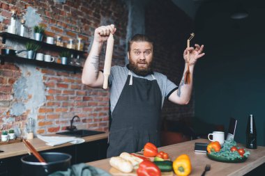 Mutfakta yemek hazırlarken tezgahın yanında merdane ve spatula tutan önlüklü belirsiz sakallı adam.