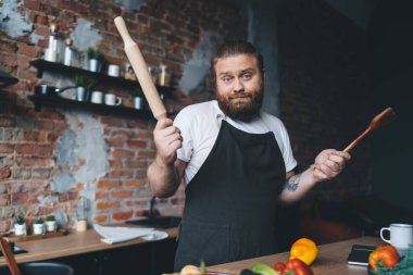 Günümüz mutfağında öğle yemeği hazırlarken elinde ahşap spatula ve merdane ile tezgahın yanında duran önlüklü cahil sakallı adam.