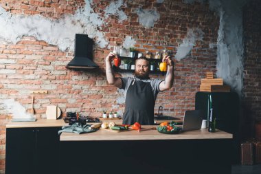 Siyah önlüklü, etkileyici sakallı bir erkek elinde dizüstü bilgisayarla, elinde kırmızı ve sarı biberle çatı katı tarzı mutfakta yemek pişiriyor.