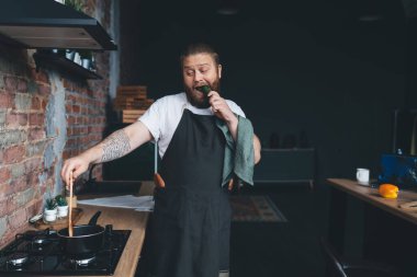 Komik ve cüsseli bir adam salatalık ısırıyor ve evde yemek pişirirken tencerede yemek pişiriyor.