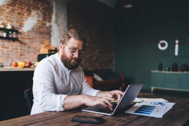 Ciddi bir erkek girişimci beyaz gömlek giyer masada oturur ve netbook 'a yazar kameraya bakar ve evdeki iş projesinde çalışır.