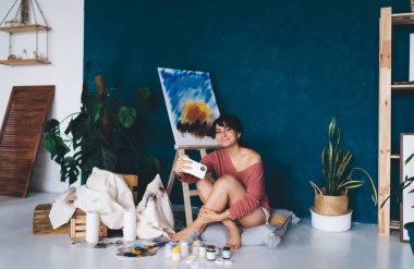 Tasasız genç bayan sanatçı resim sehpası yakınında akıllı telefon kamerası için poz veriyor ve blogunda hobisini gösteren resimler yapıyor. Yaratıcı gülümseyen esmer kadın, yağlı boya resminin yanında kendini fotoğraflıyor. 