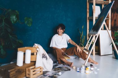 Heyecanlı genç bayan sanatçı stüdyo zemininde oturmuş, resimleriyle hobisinin tadını çıkarıyor. Yaratıcı ve yetenekli esmer kadın sehpada resim yaratıyor. Petrolle çizim yapmayı öğrenmek