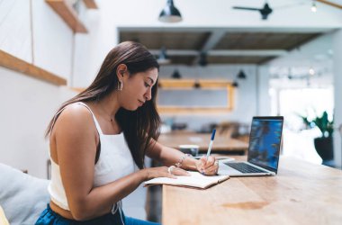 Genç İspanyol kadın dizüstü bilgisayarda çalışıyor, birlikte çalışıyor, not defterine notlar yazıyor. İnternette serbest çalışan kadın internet sitesi. Öğrenci webinar öğreniyor, bilgi okuyor