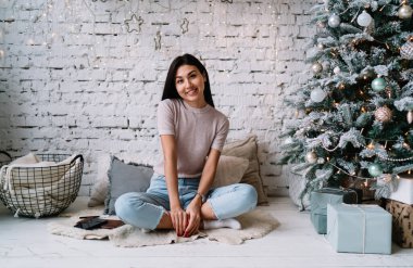 Gülümseyen Çinli kız kürklü halıya oturur ve Noel ağacının yanındaki kameraya bakar evde hediyelerle. Ev eğlencesi ve eğlence. Noel ve Yeni Yıl. Milenyum kuşağının genç kadını