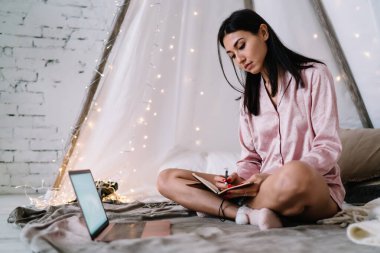 Genç Asyalı kadın evde dizüstü bilgisayar ve çelenklerle çadırda otururken deftere bir şeyler yazıyor. Eğlence ve eğlence anlayışı. Pijama giyen esmer kız. Noel ve Yeni Yıl Zamanı