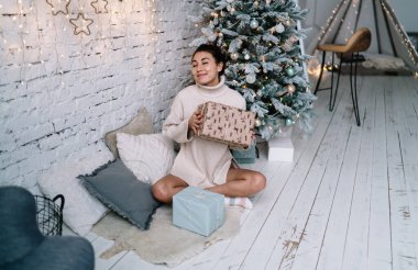 Tüylü halıda oturan ve Noel ağacının yanında hediyesini tutan mutlu Çinli kız. Noel ve yeni yıl tatilleri. Gözleri kapalı genç bir kadın süveter giyer. Ev hayatı işte. Modern daire