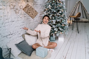 Neşeli Çinli kız, evdeki Noel ağacının yanında kürklü halıda otururken hediye kutusunu kusuyor. Noel ve yeni yıl tatilleri. Süveter giyen genç bir kadın. Ev hayatı işte. Geniş daire