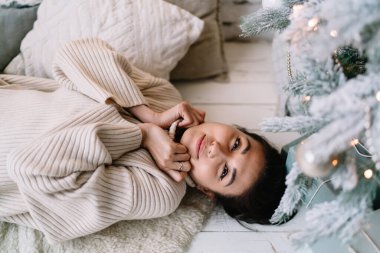Çinli kızın tüylü halıda yatıp evdeki Noel ağacının yanındaki kameraya bakarken kısmi görüntüsü. Noel ve yeni yıl tatilleri. Genç esmer kadın kazak giyiyor. Yurt içi yaşam tarzı