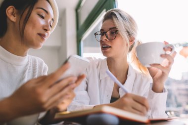 Kafe içi, Asyalı ve Kafkasyalı kadınlar canlı ve sosyal ağ hakkında iletişim kurarken cep telefonlu çekici kadın blogcular, içerik fikirlerini tartışıyorlar.